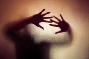 Colour backlit image of the silhouette of a woman with her hands pressed against a glass window. The silhouette is distorted, and the arms elongated, giving an alien-like quality. The image is sinister and foreboding, with an element of horror. It is as if the 'woman' is trying to escape from behind the glass. Horizontal image with copy space. Dark Hollow